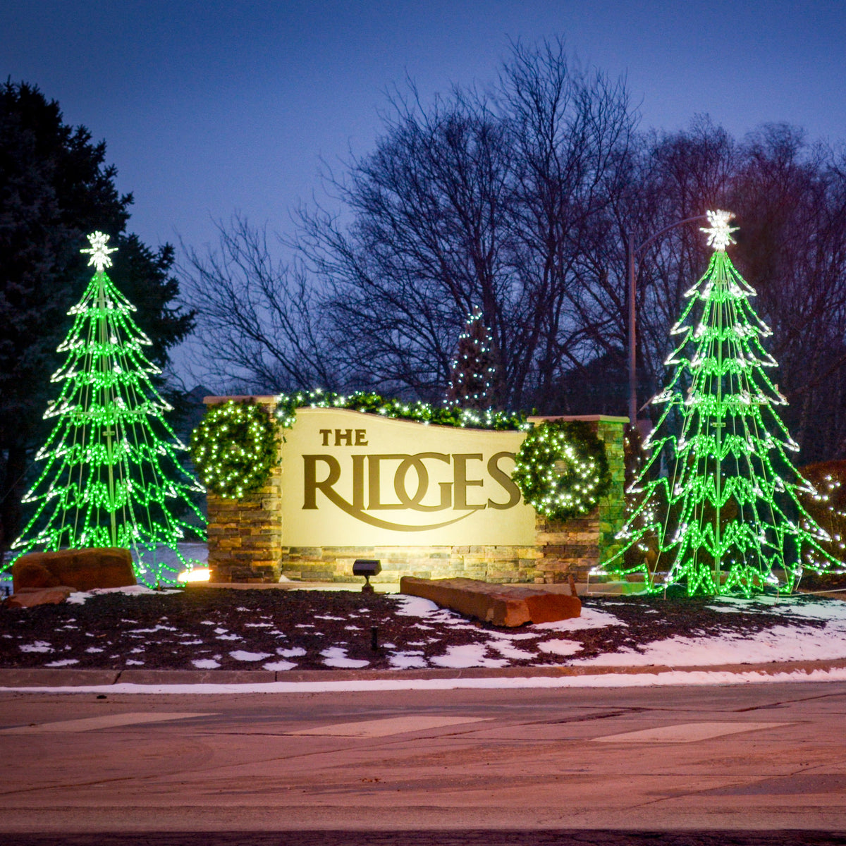 Two LED 3D Trees 10 ft. flanking a stone community entrance sign at dusk, green and white LED layered cone silhouettes with starburst star toppers installed in snow-dusted landscape beds